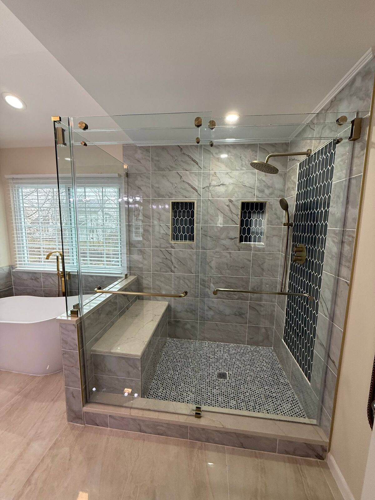 A large frameless glass shower enclosure with brass hardware, a tiled bench and a freestanding tub beside it