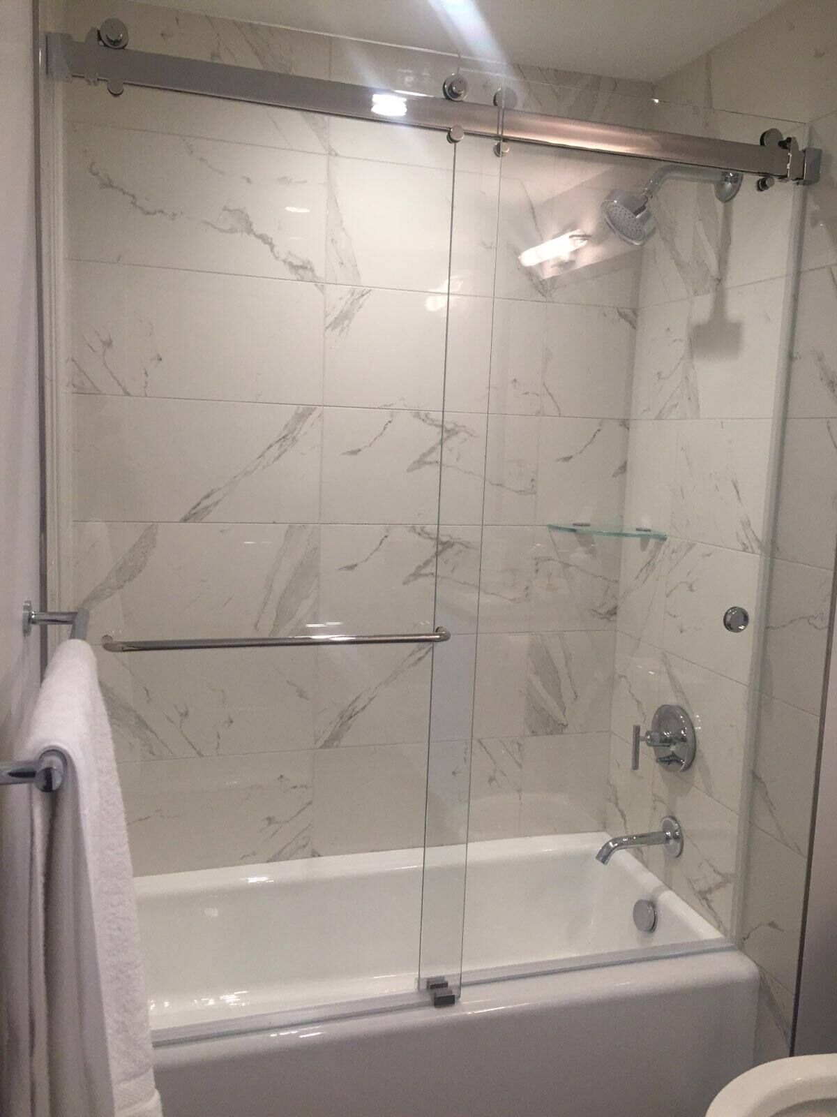 Frameless sliding glass doors on a top roller rail over a white bathtub with marble-look tile