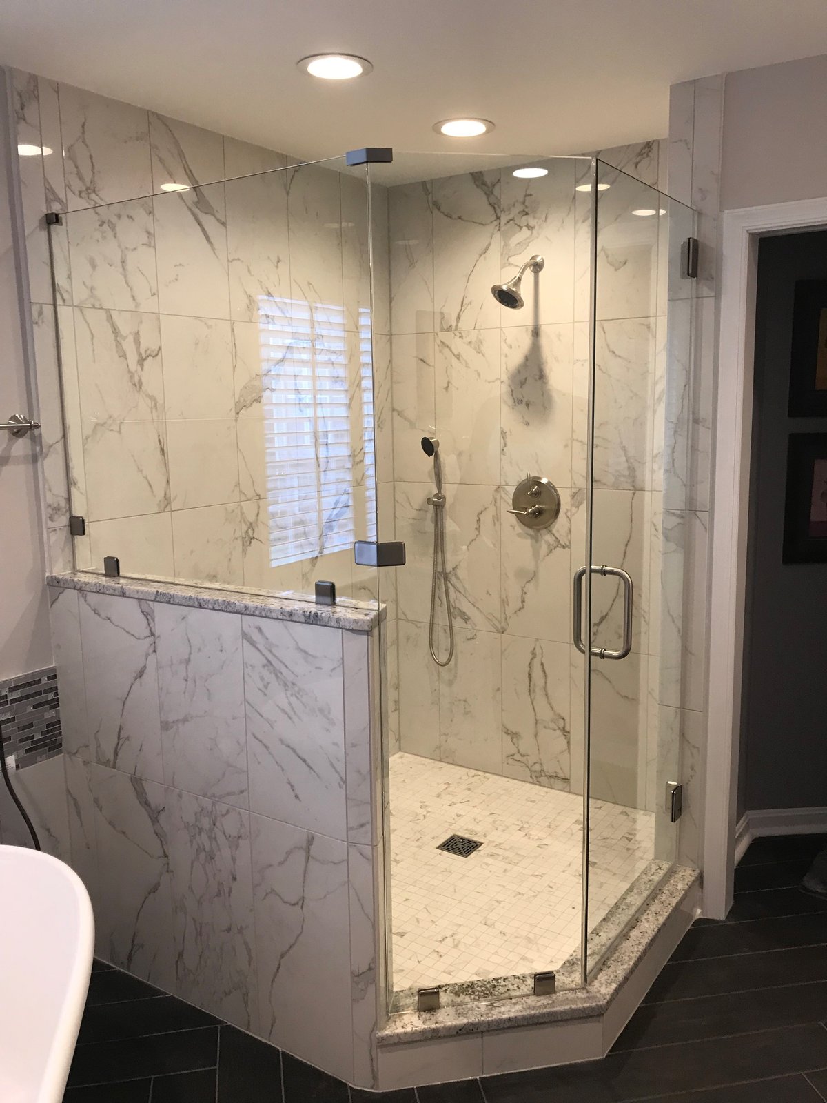 A frameless neo-angle shower with one glass panel set on a marble-look tiled half wall